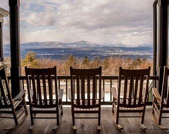 May include: Four wooden rocking chairs are arranged in a row on a porch, facing a panoramic view of snow-dusted mountains and a forest under a cloudy sky. The chairs feature vertical slats and curved rockers, positioned on a wooden deck floor.