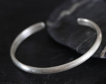 Puede incluir: Un brazalete simple de plata con un acabado liso y pulido. El brazalete está abierto y se encuentra sobre una superficie gris oscura.