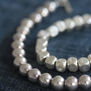 Puede incluir: Dos pulseras de plata con cuentas redondas y cuadradas. Las pulseras están sobre un fondo de tela azul.