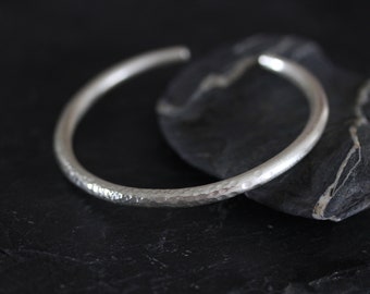 Puede incluir: Un brazalete de plata martillado. El brazalete es liso y brillante, con una superficie ligeramente texturizada.