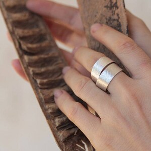 May include: A silver ring with a hammered texture, worn on a finger. The ring is made of two bands that are wrapped around each other.