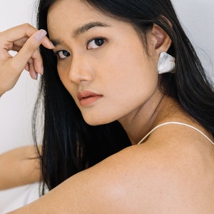 Puede incluir: Una mujer con el pelo largo y negro que lleva un top blanco sin mangas y pendientes de plata. Está mirando a la cámara con una expresión seria.