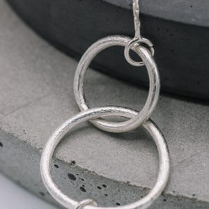 May include: A silver necklace with two interlocking circles. The circles are textured and have a brushed finish. The necklace is on a gray background.