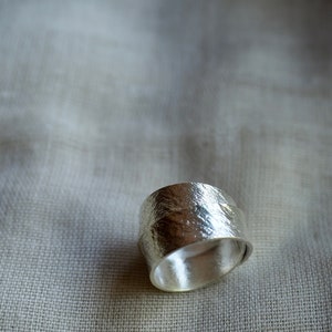 May include: A silver ring with a textured, hammered finish. The ring is wide and has a rounded shape.