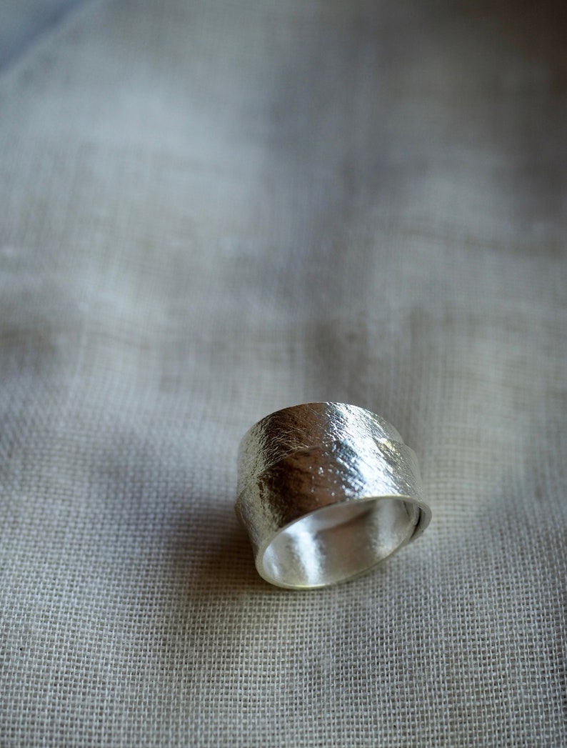 May include: A silver ring with a textured, hammered finish. The ring is wide and has a rounded shape.