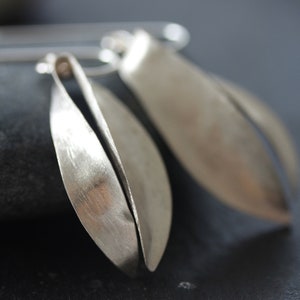 Puede incluir: Pendientes plateados con forma de hoja. Los pendientes tienen un acabado de metal cepillado y un dise&ntilde;o abierto &uacute;nico. Las formas alargadas y curvas crean una est&eacute;tica moderna y minimalista. Los pendientes se muestran sobre un fondo oscuro.