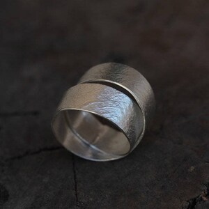May include: A silver ring with a unique, twisted design. The ring is made of two bands that are intertwined and have a textured finish.