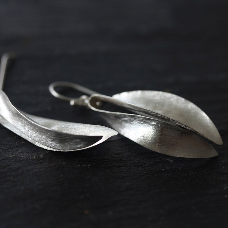 Puede incluir: Un par de pendientes plateados con forma de hoja. Los pendientes tienen una superficie texturizada y un cierre de gancho. El dise&ntilde;o es minimalista y elegante, centrado en la forma natural de las hojas. Se muestran sobre un fondo oscuro.