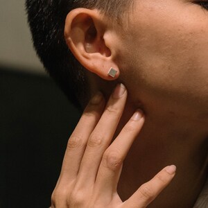 May include: A person wearing a small, square silver earring. The earring is in the person's left ear.