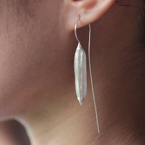 May include: Silver dangle earrings featuring a long, thin, silver rod with a small, textured, silver rice grain shape at the bottom.