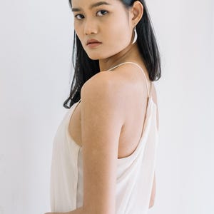 Puede incluir: Una mujer con cabello negro lleva una camiseta sin mangas color crema y pendientes plateados en forma de media luna. El fondo es una pared blanca lisa. Los pendientes son una caracter&iacute;stica destacada.
