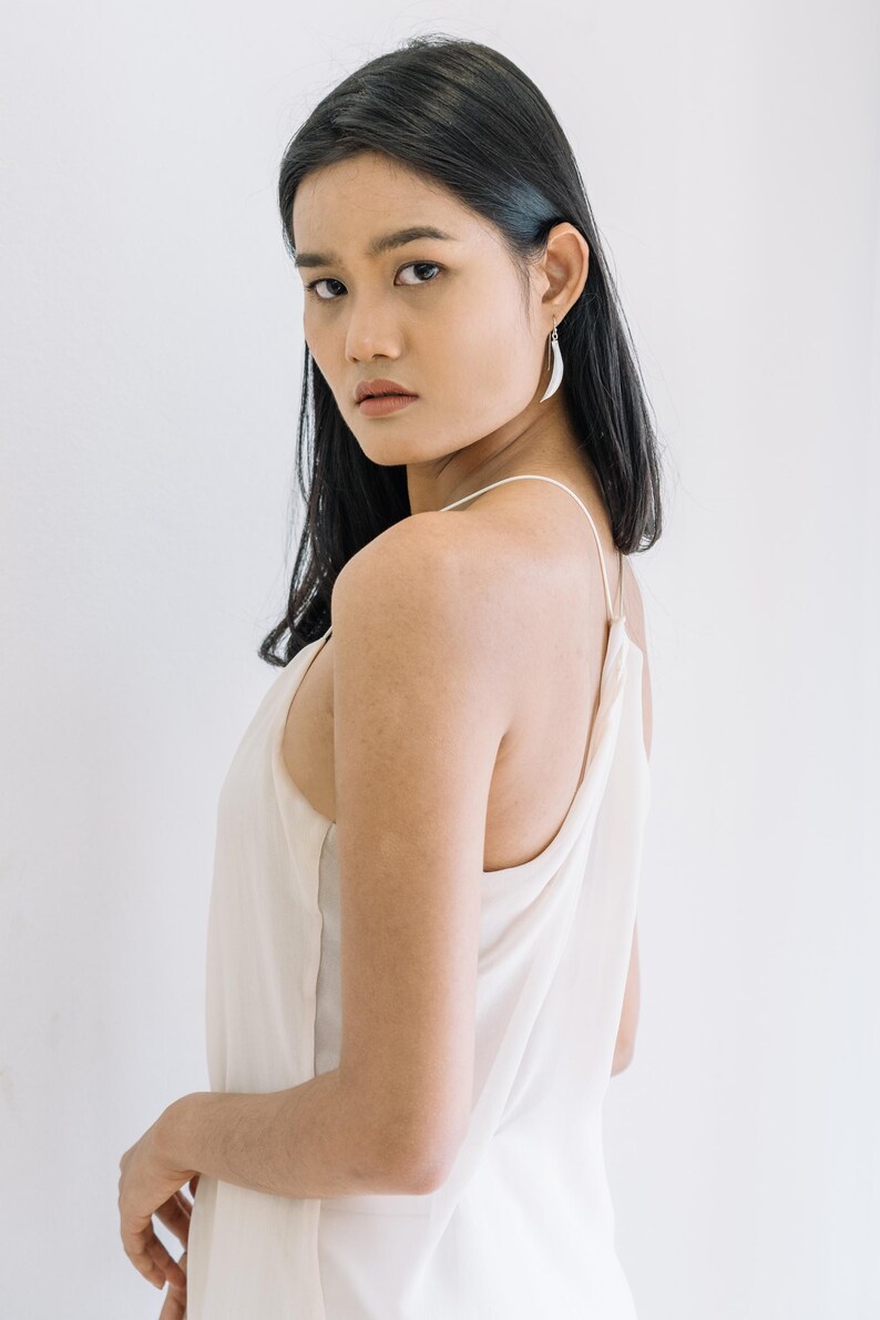 Puede incluir: Una mujer con cabello negro lleva una camiseta sin mangas color crema y pendientes plateados en forma de media luna. El fondo es una pared blanca lisa. Los pendientes son una caracter&iacute;stica destacada.