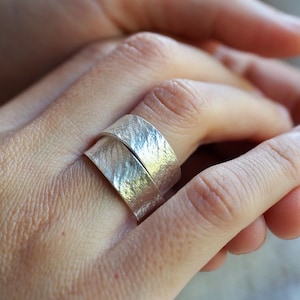 May include: A silver ring with a textured finish. The ring is worn on a person's finger.