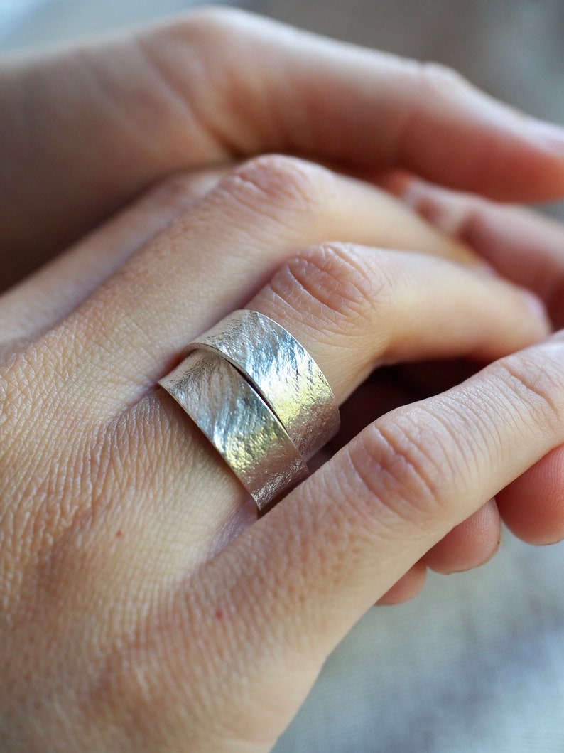 May include: A silver ring with a textured finish. The ring is worn on a person's finger.