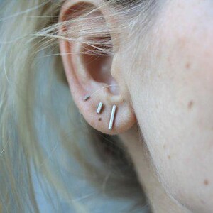 May include: A close-up of a person's ear with three small silver bar earrings. The earrings are in the helix, lobe, and upper lobe piercings.
