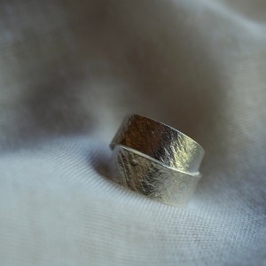 May include: A silver ring with a textured, hammered finish. The ring is designed to be worn on the finger with the band wrapping around twice.