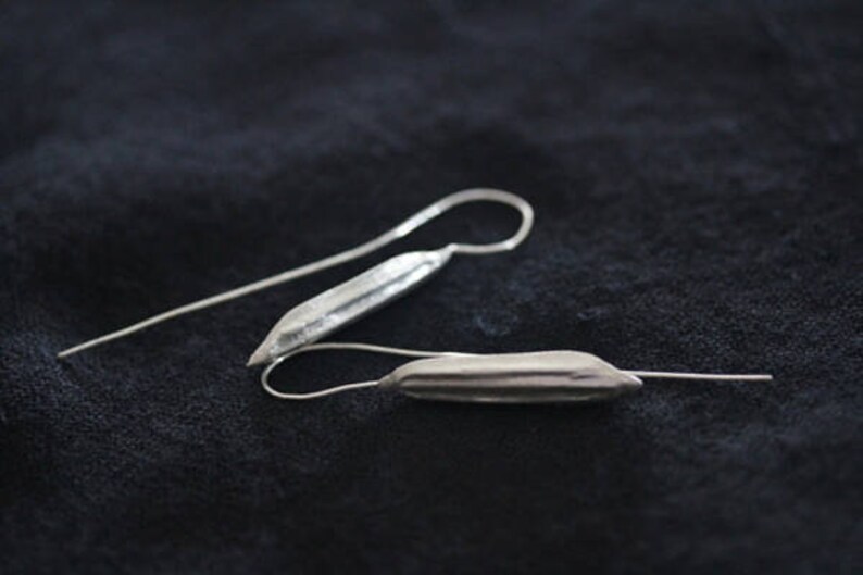 May include: A pair of silver dangle earrings with a long, thin, and slightly curved design. The earrings have a textured, ridged surface.