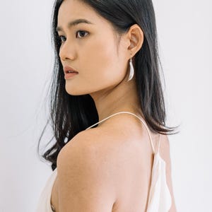Puede incluir: Una mujer con pendientes colgantes plateados. Los pendientes tienen un dise&ntilde;o curvo en forma de hoja. Lleva una camiseta blanca de tirantes finos. Su cabello oscuro es largo y ondulado. El fondo es una pared blanca lisa.