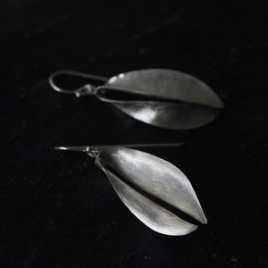 Puede incluir: Un par de pendientes plateados con dise&ntilde;o de hoja. Cada pendiente presenta una forma curva y alargada con una hendidura en el centro y un cierre de gancho. Los pendientes est&aacute;n sobre un fondo oscuro, resaltando su brillo met&aacute;lico.