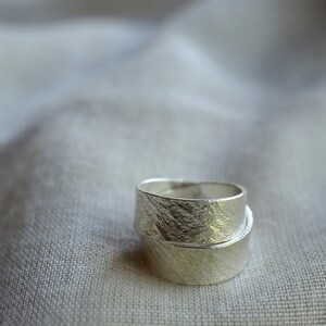 May include: Two silver rings with a textured finish, stacked on top of each other. The rings are on a white fabric background.