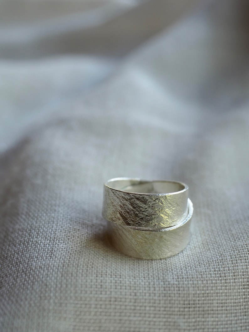 May include: Two silver rings with a textured finish, stacked on top of each other. The rings are on a white fabric background.
