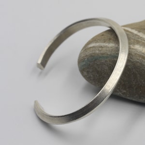 May include: A silver bangle bracelet with a smooth, rounded finish. The bracelet is resting on a gray rock.