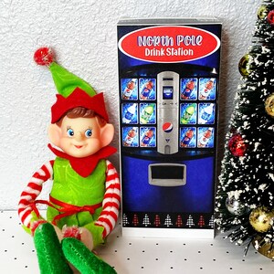 May include: A green elf doll with red and white striped sleeves sits in front of a blue and silver vending machine with the text "North Pole Drink Station". The vending machine has a variety of drinks, including soda, water, and juice. The elf is sitting on a white surface with black polka dots.