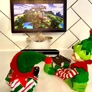 May include: Two elf figurines playing a video game on a computer monitor. The monitor displays the game Minecraft. The elves are wearing green and red striped outfits and Santa hats. One elf is holding a video game controller.