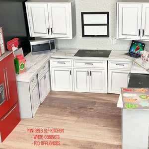 May include: A miniature dollhouse kitchen with white cabinets and red appliances. The kitchen features a microwave, stovetop, refrigerator, and a countertop with a cookie sheet and Christmas-themed cookie cutters. The text "Printable Elf Kitchen - White Cabinets - Red Appliances" is printed below the kitchen.