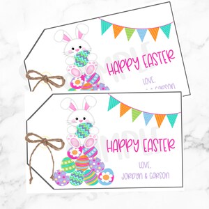 Könnte beinhalten: Weiße Oster-Geschenkanhänger mit einem Cartoon-Hasen, der ein gemustertes Ei hält. Die Anhänger haben ein farbenfrohes Banner, die Worte "HAPPY EASTER" und Platz für einen Namen. Eine Schnur-Schleife ist befestigt.