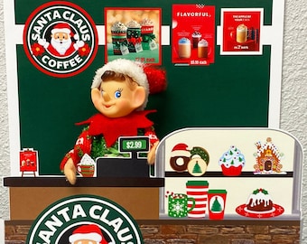 May include: An elf wearing a green and red sweater stands behind a counter at a Santa Claus Coffee shop. The counter has a cash register with a price tag of $2.99. There are various Christmas-themed drinks and pastries on display behind the counter.