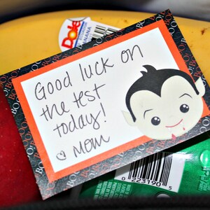 May include: A Halloween-themed note with a vampire illustration and the text "Good luck on the test today! Love Mom". The note is on a black and orange background with a white border. The note is sitting on top of a lunch bag with a red apple, two yellow bananas, and a green yogurt container.