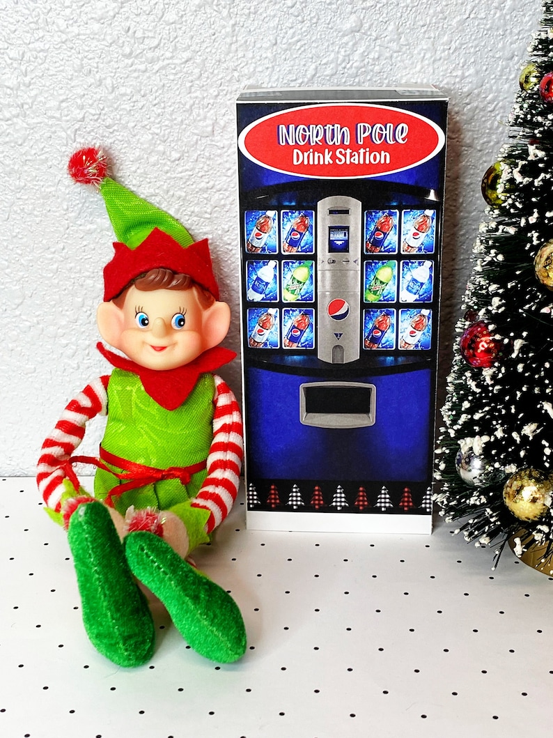 May include: A green elf doll with red and white striped sleeves sits in front of a blue and silver vending machine with the text "North Pole Drink Station". The vending machine has a variety of drinks, including soda, water, and juice. The elf is sitting on a white surface with black polka dots.