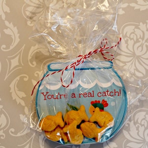 May include: A blue and white fishbowl-shaped card with the text "You're a real catch!" and a red and white string bow. The card is filled with goldfish crackers.