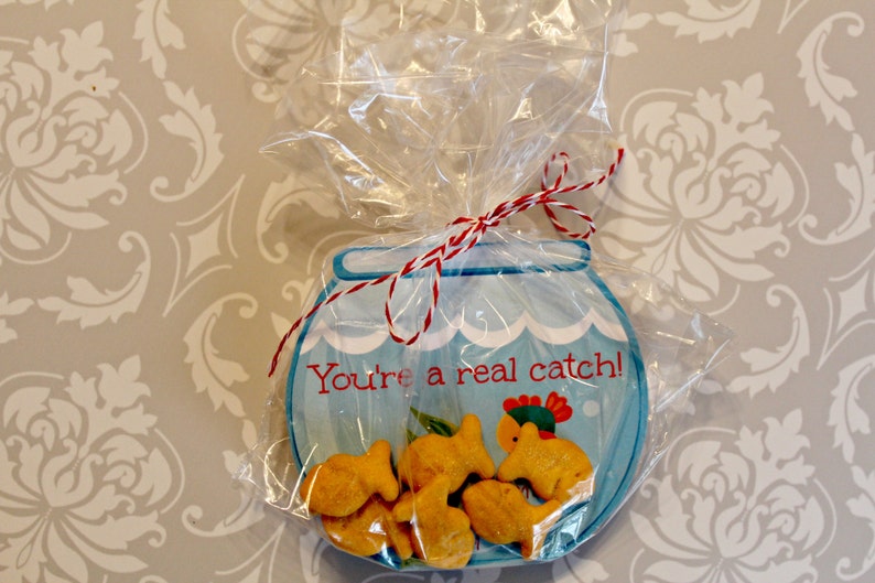 May include: A blue and white fishbowl-shaped card with the text "You're a real catch!" and a red and white string bow. The card is filled with goldfish crackers.