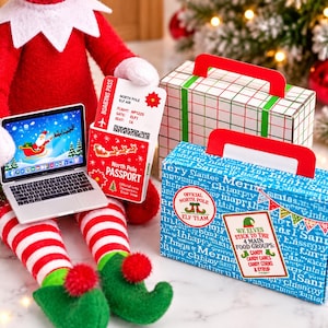Puede incluir: Un muñeco elfo de peluche con piernas a rayas rojas y blancas y zapatos verdes sostiene un portátil en miniatura con Santa en un trineo, un pasaporte del Polo Norte y una tarjeta de embarque. Hay dos cajas de regalo.