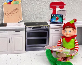 May include: A green elf doll with red and white striped sleeves is holding a baking sheet with cookies. The elf is sitting on a white floor with black polka dots. The doll has a red hat and a green shirt. The baking sheet has a gingerbread man, a snowman, a wreath, and a star cookie on it.