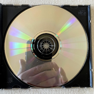 May include: A circular silver compact disc with a reflective surface showing rainbow light refractions, housed in a black plastic case. The disc features a central black hub and the text Compact Disc Digital Audio printed in the upper right corner.