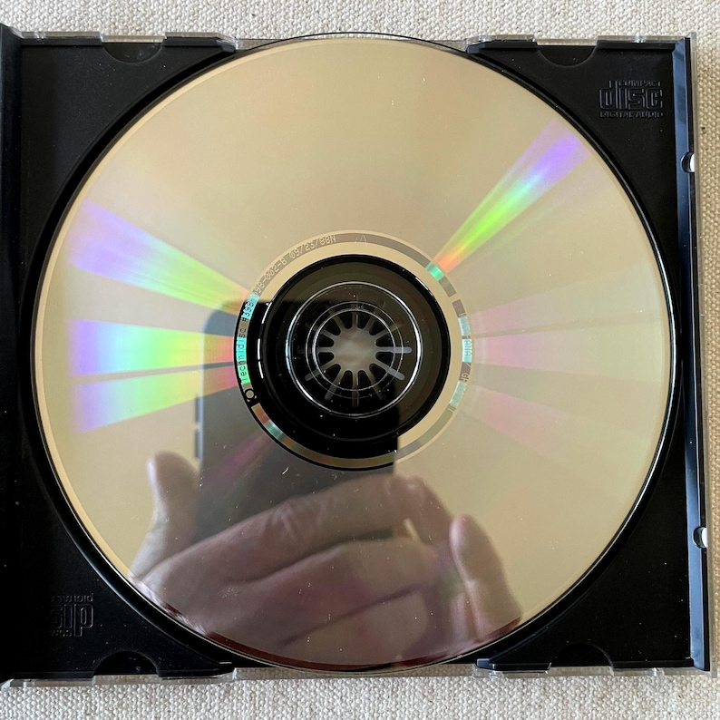 May include: A circular silver compact disc with a reflective surface showing rainbow light refractions, housed in a black plastic case. The disc features a central black hub and the text Compact Disc Digital Audio printed in the upper right corner.