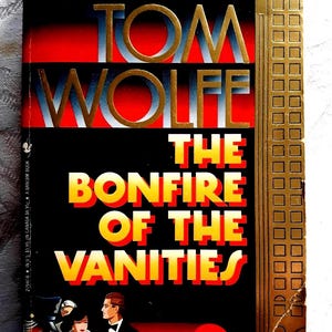以下が含まれることがあります： トム・ウルフ著『The Bonfire of the Vanities』のペーパーバック。表紙は黒い背景に赤と黄色の文字、右側に金色の高層ビルのイラスト、手前に白いリムジンと人物が描かれています。