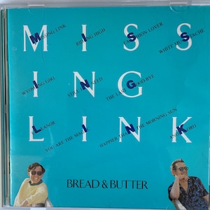 Puede incluir: Una caja de CD azul con el título "Missing Link" en letras blancas. La caja presenta un collage de títulos de canciones del álbum, incluyendo "Riding High", "Wyoming Girl", "Still I Need", "The Last Good-Bye", "Happier Than The Morning Sun", y "Ano Koro". La caja también presenta dos fotos de personas, una a cada lado.