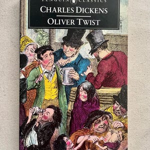 Puede incluir: Una edición de Penguin Clásicos de "Oliver Twist" de Charles Dickens. La portada presenta una colorida ilustración de una multitud con vestimenta de época. El título y el nombre del autor están en blanco sobre una pancarta negra.