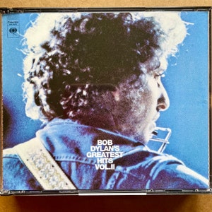 May include: A blue and white CD case featuring a photograph of Bob Dylan on the front. The text on the case reads "Bob Dylan's Greatest Hits Vol. II".