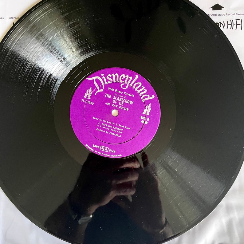 May include: A vintage black vinyl record with a purple label featuring the text "Disneyland" and "The Scarecrow of Oz" with Ray Bolger. The label also includes the song title "Over the Rainbow". The record is a 33 1/3 RPM long play.