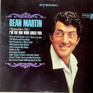 May include: A vinyl record sleeve featuring a portrait of Dean Martin on the right. The left side displays the text Dean Martin, Remember Me, I'm The One Who Loves You, and a tracklist including King of the Road, all in white and blue lettering.