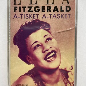Puede incluir: Un casete vintage del álbum "A-Tisket, A-Tasket" de Ella Fitzgerald. La portada muestra un retrato en tonos sepia de Fitzgerald, con el título del álbum en letras negras. El casete está en una caja de plástico transparente.