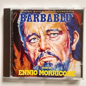 May include: A CD case featuring the "Barbablu" original motion picture soundtrack. The cover art showcases a painted portrait of a man with blue eyes and a beard. The text "Barbablu" and "Music by Ennio Morricone" are also visible.