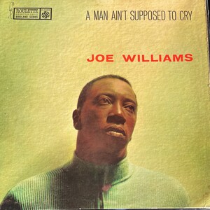 May include: A vintage vinyl record album cover featuring a black and white photograph of a man with a serious expression. The album title is "A Man Ain't Supposed To Cry" and the artist is "Joe Williams". The album is part of the "Roulette Birdland Series".