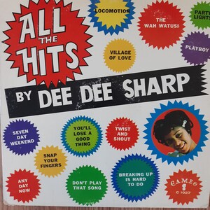 May include: A white vinyl record album cover with a red starburst design featuring the text "All the Hits by Dee Dee Sharp". The album cover features a variety of song titles in colorful starburst designs, including "Seven Day Weekend", "You'll Lose a Good Thing", "Twist and Shout", "Snap Your Fingers", "Don't Play That Song", "Breaking Up is Hard to Do", "Any Day Now", "Locomotion", "Village of Love", "The Wah Watusi", "Party Lights", and "Playboy". The album cover also features a photo of Dee Dee Sharp in a colorful starburst design.