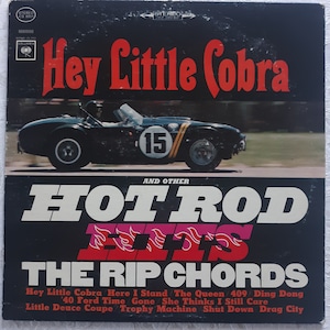 May include: A vintage vinyl record album cover featuring a black Cobra sports car with the number 15 on the side, racing on a track. The album title is "Hey Little Cobra and Other Hot Rod Hits" by The Rip Chords. The album cover is black with red and white text.